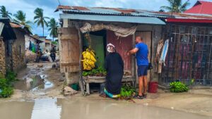 obchod s potravinami na zanzibaru,kde visí trs banánů. Evropský muž si kupuje jídlo od africké ženy.