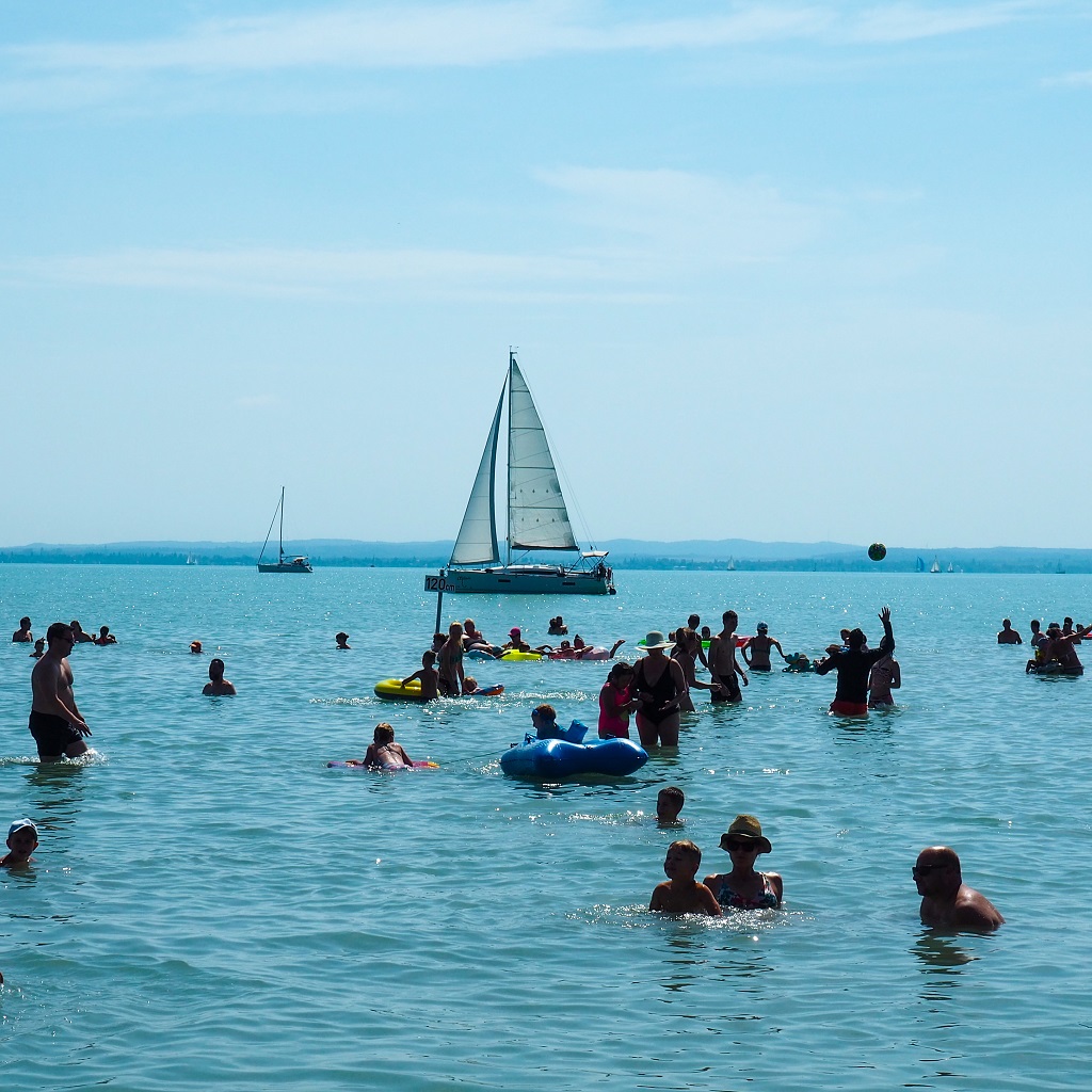 lidé se koupají ve vodě v jezeře Balaton, v pozadí pluje jachta