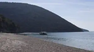 Široká oblázková pláž Krorëz s kotvící lodí a zeleným kopcem v pozadí