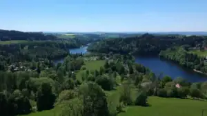 Výhled na letní krajinu v okolí Pastvinské přehrady. Zelené lesy a meandrující vodní hladina tvoří malebnou scenérii ideální pro výlety, koupání a relaxaci.